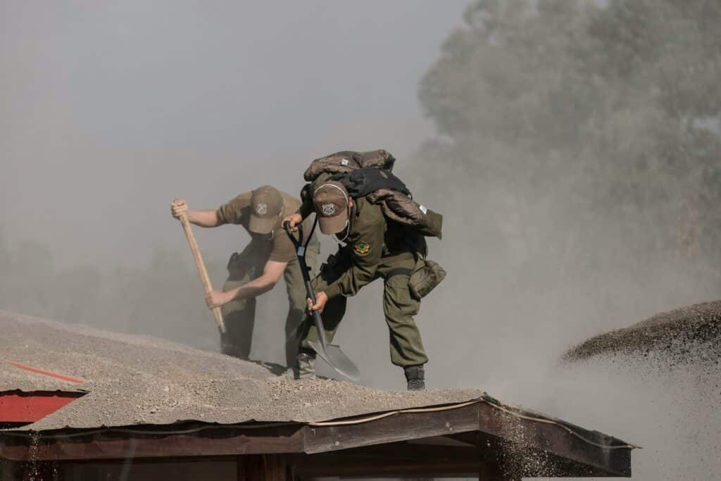 Two uniformed responders shovelling thick ash off a building roof in a cloud of grey dust during a clean-up operation.